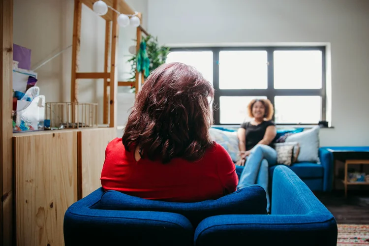 Two women sitting in a cozy living room, one with back to camera, the other smiling by a window.
