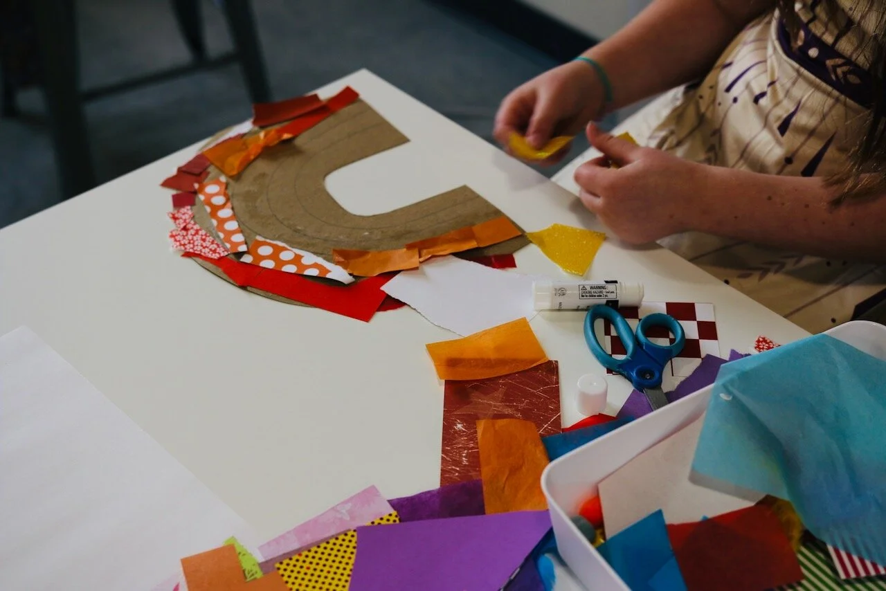 Child assembling colorful paper pieces on cardboard for a craft project on a white table.