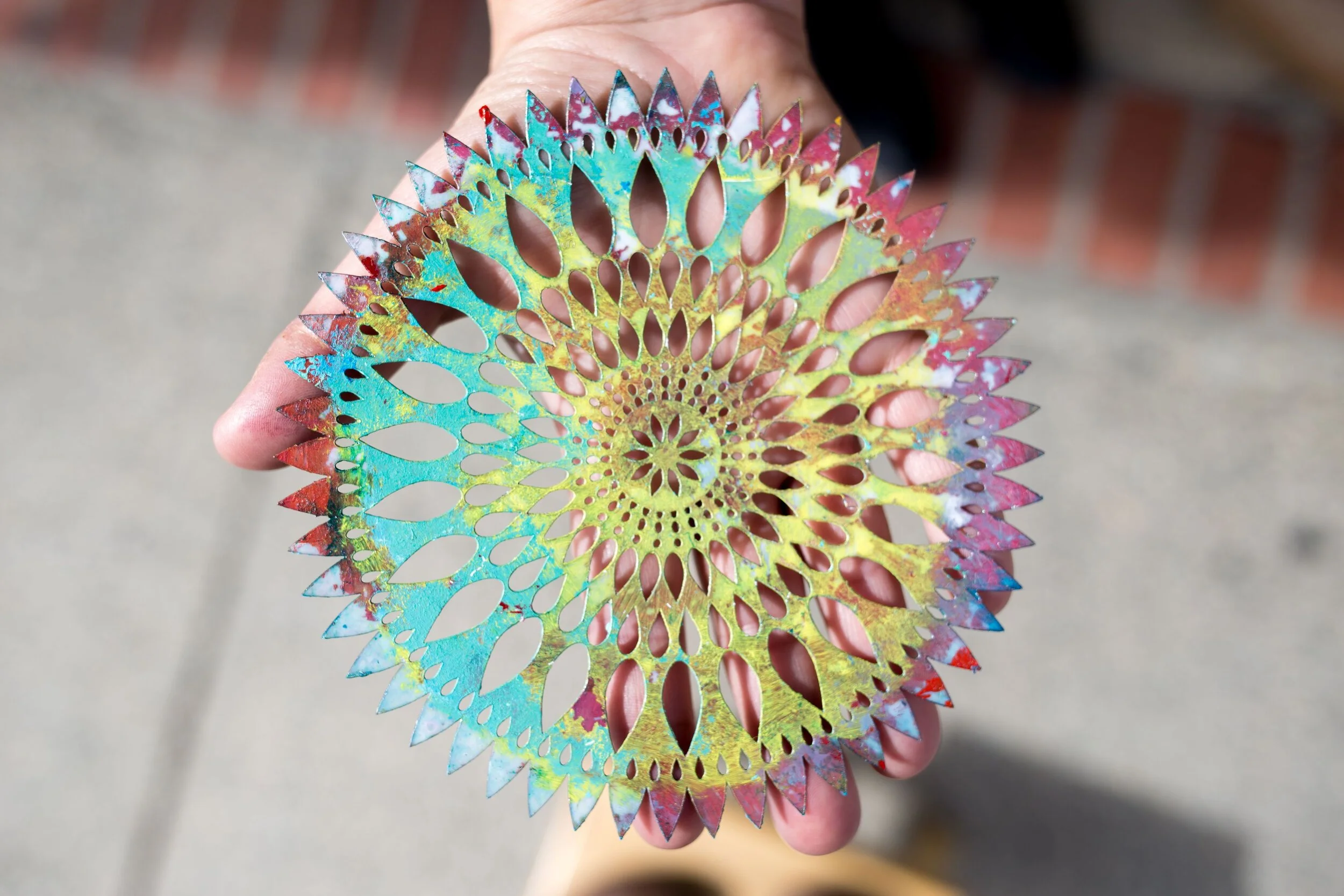 Hand holding a colorful, round, perforated metal artwork with intricate cut-out patterns.