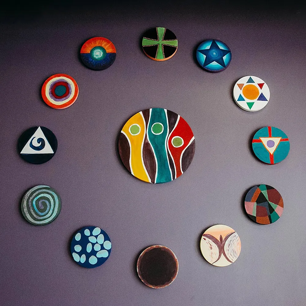 Twelve round disks painted with colorful abstract and geometric designs arranged in a circle on a purple wall.