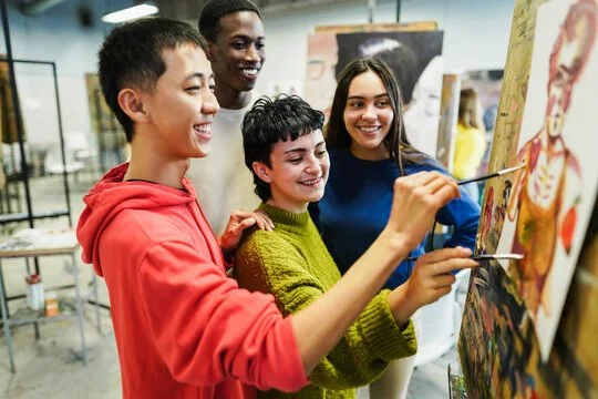 Four diverse teens smiling and painting together on a canvas in an art studio.