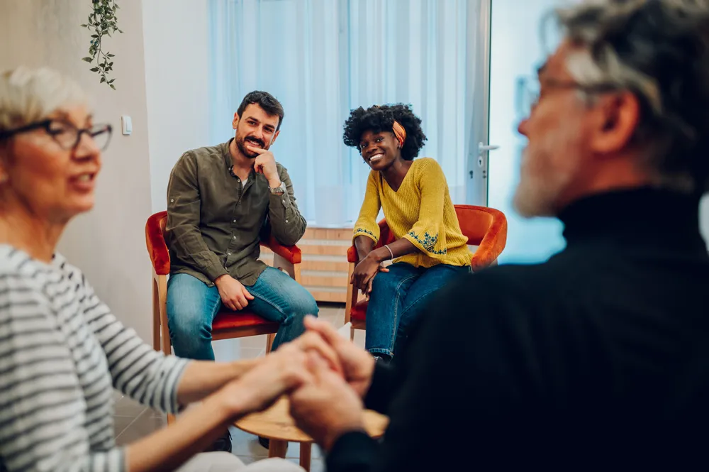 Four people in a support group meeting, two holding hands in foreground and two smiling in background