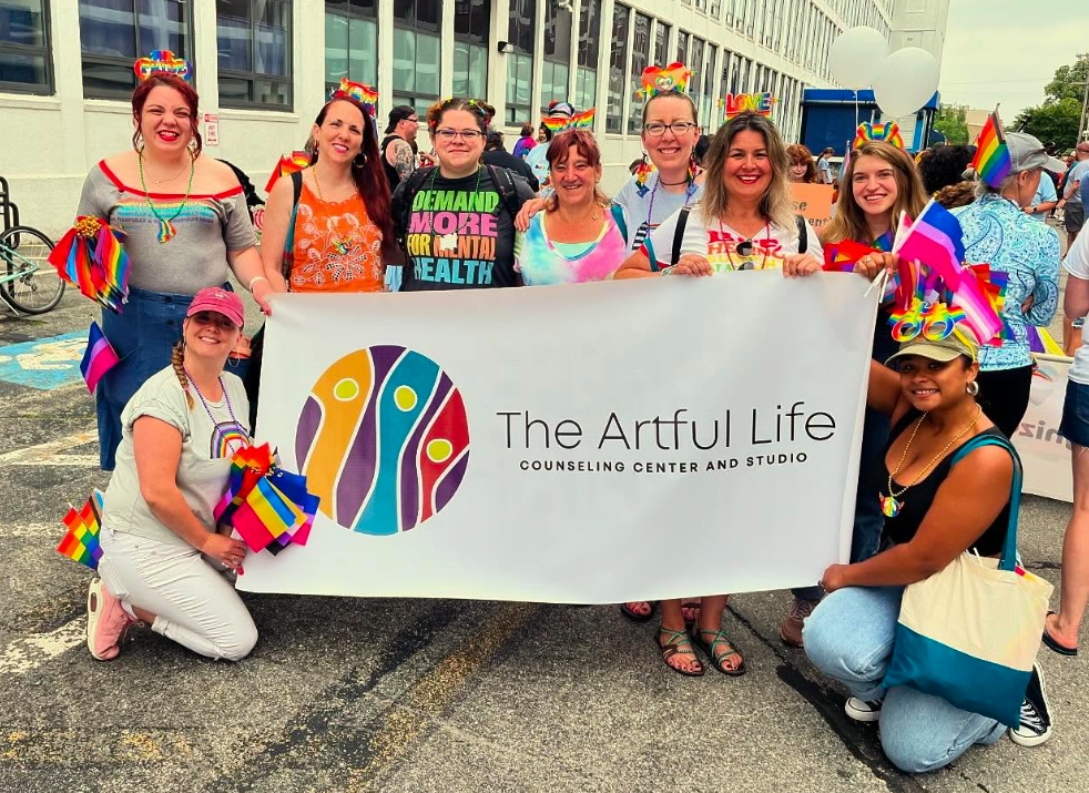 Group holding The Artful Life banner at pride event, displaying rainbow and LGBTQ+ flags and smiles.