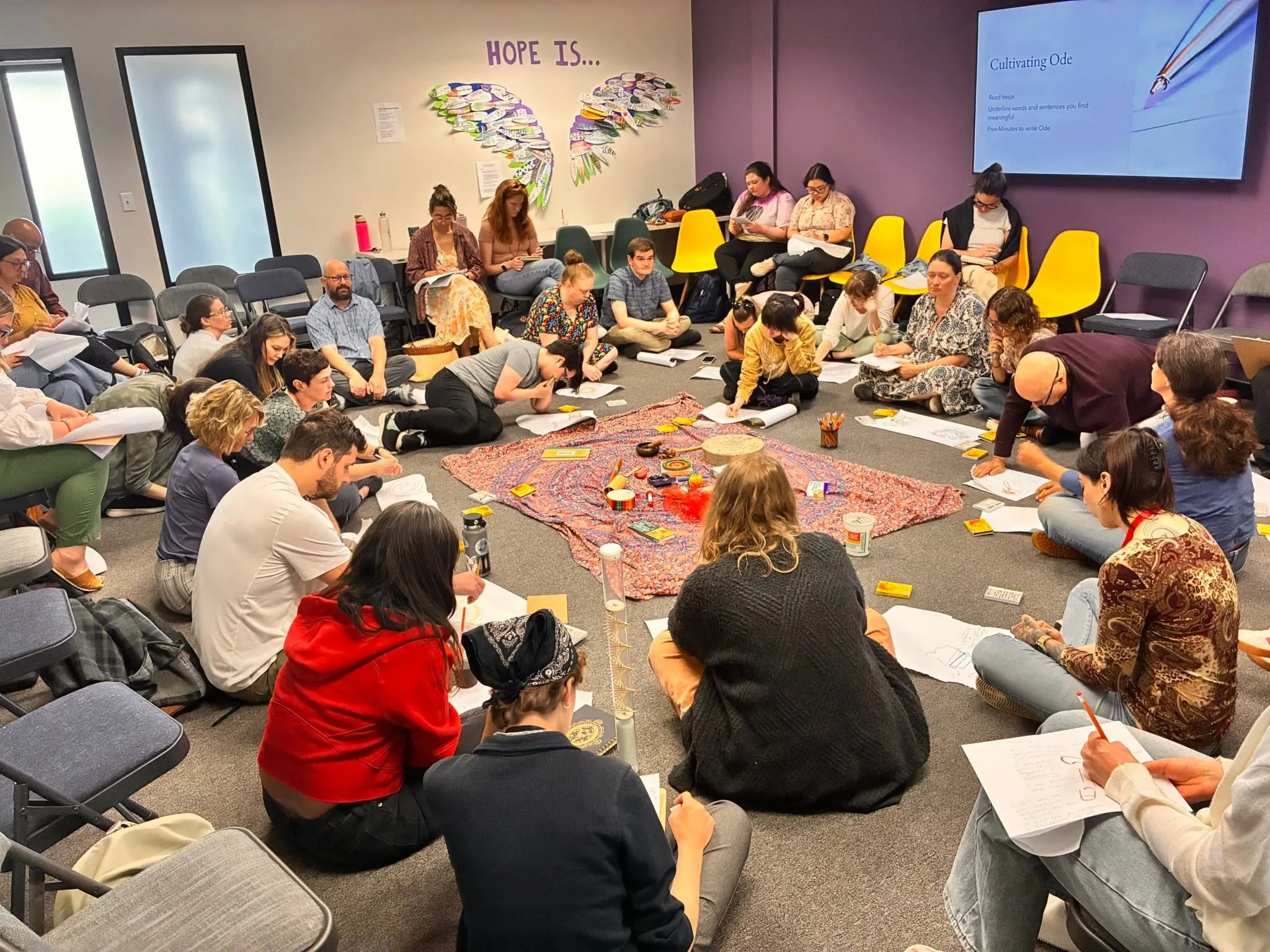 Group of people sitting in a circle on floor, writing on paper around art supplies and fabrics.