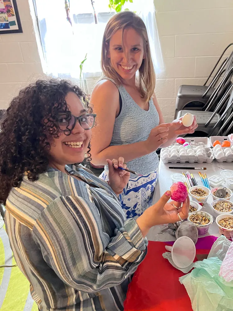 Two smiling women painting eggs at a table with art supplies and paintbrushes.