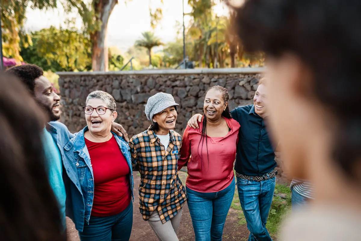 Smiling multigenerational group standing arm in arm outdoors with greenery and stone wall behind them.
