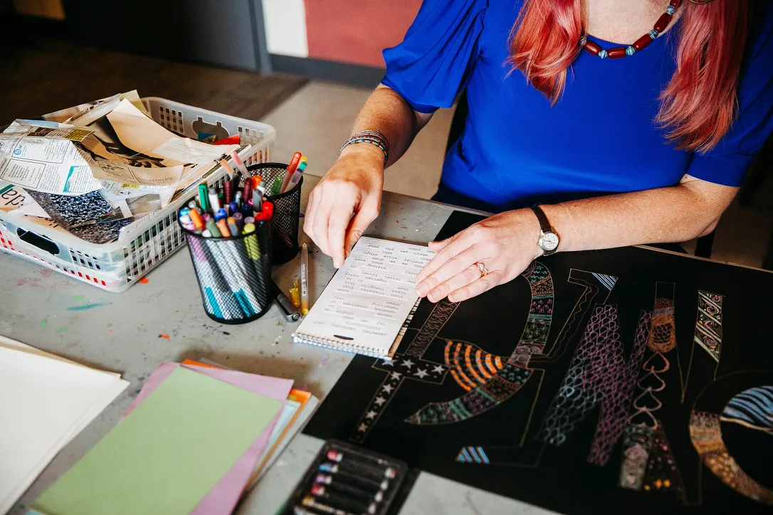 Person with pink hair and blue shirt doing colorful paper art on a table with art supplies.