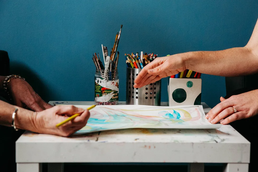 Two people using paintbrushes and colored pencils to create art on paper at a white table.