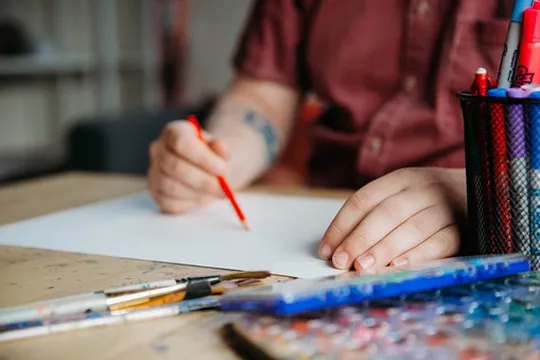 Person holding a red colored pencil over blank paper with art supplies on wooden table.