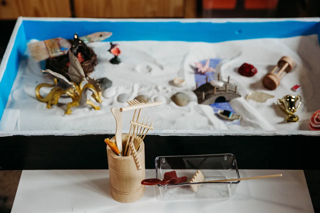 A tabletop sand tray with miniature objects and a container holding wooden tools for sand play therapy.