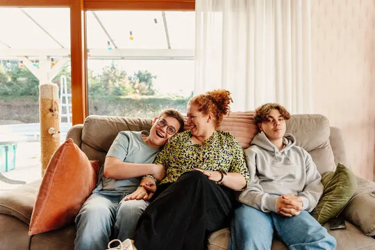 Woman with two teenage boys sitting together on a couch in a sunlit room near a window.
