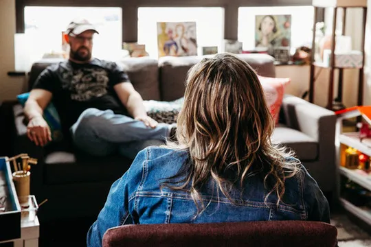 Man in a cap sitting on a couch facing a woman with long hair in a denim jacket indoors.