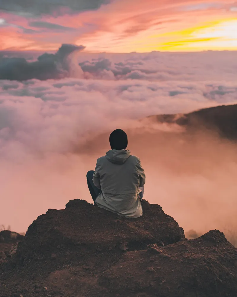 Person sitting on rocky cliff above clouds watching colorful sunset sky.