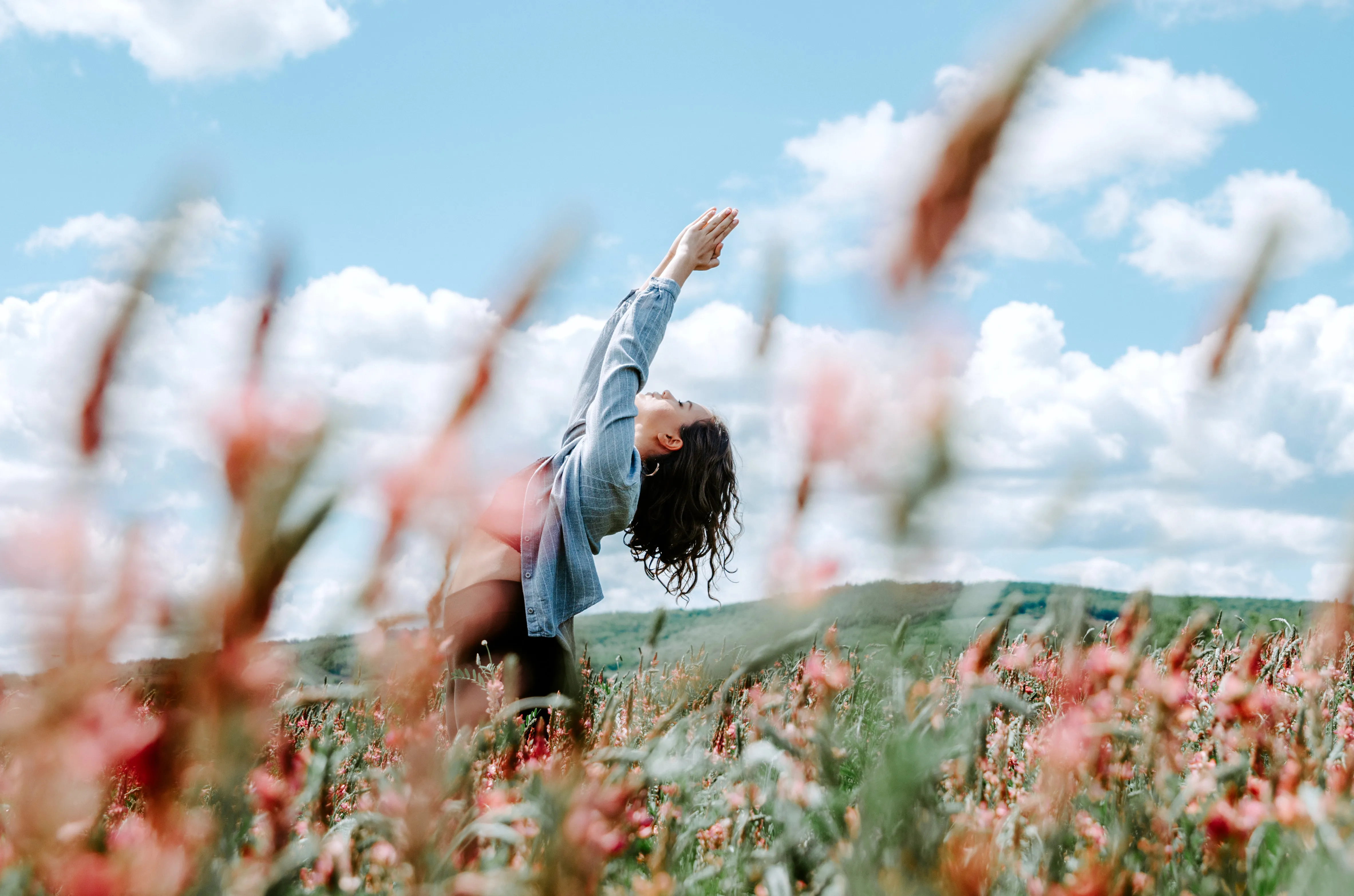 Young woman stretching with hands raised leaning back in a flower field under blue sky with clouds.