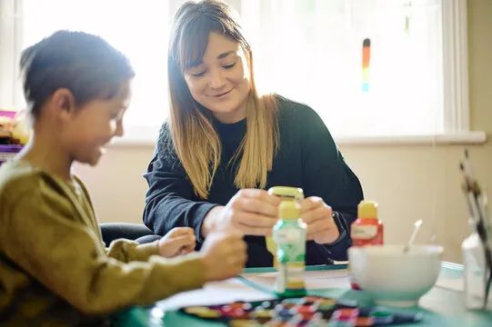 Woman and child painting together at a table with art supplies and paint bottles.