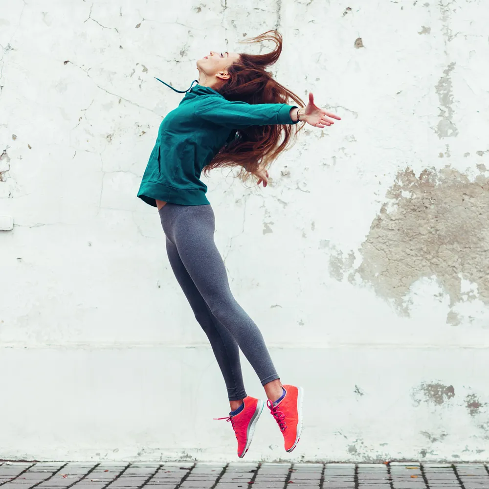 Young woman in teal jacket and gray leggings jumping with arms wide against a cracked wall.