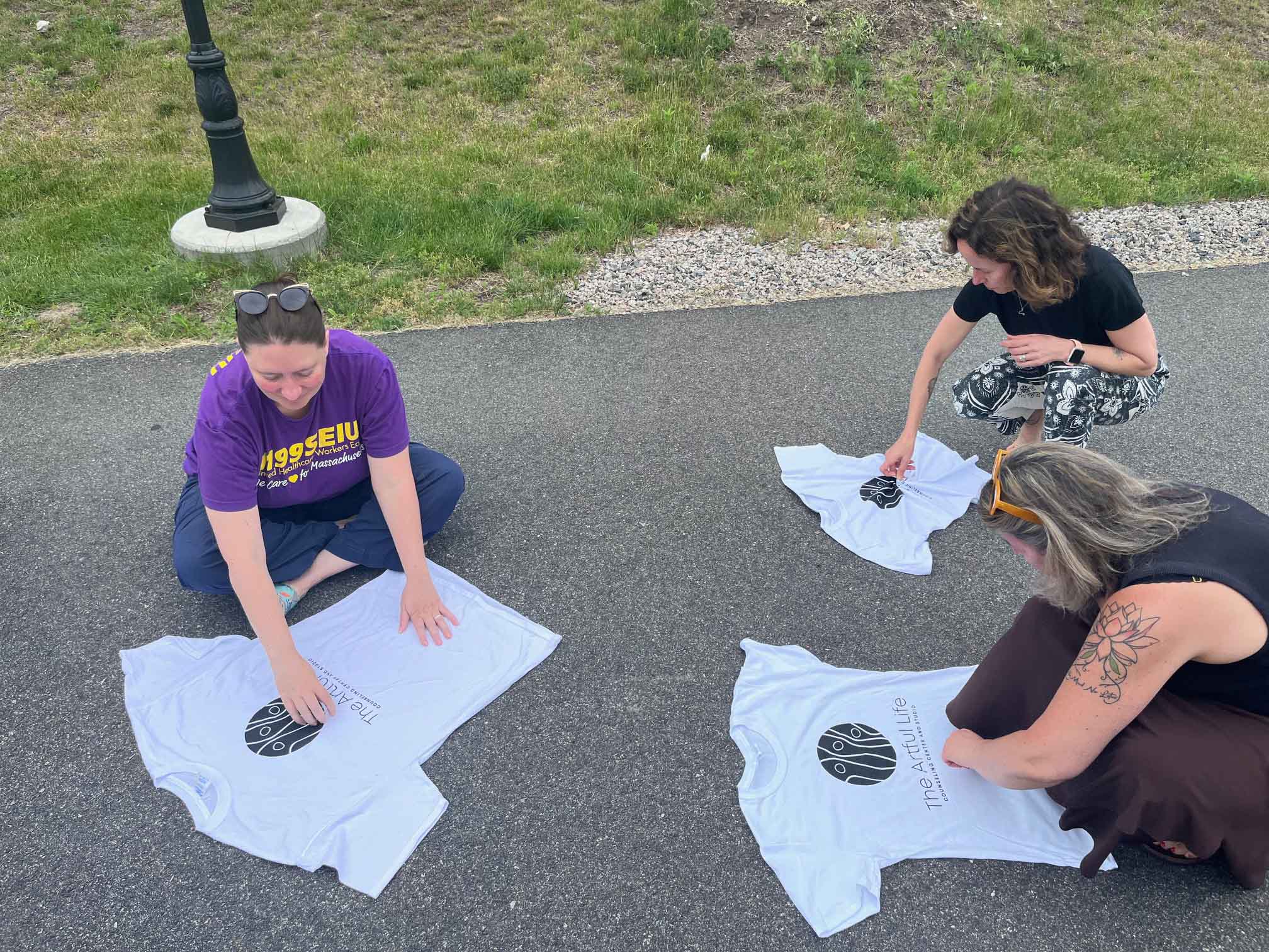 Three people sitting on asphalt outdoors arranging white T-shirts with a black circular logo.