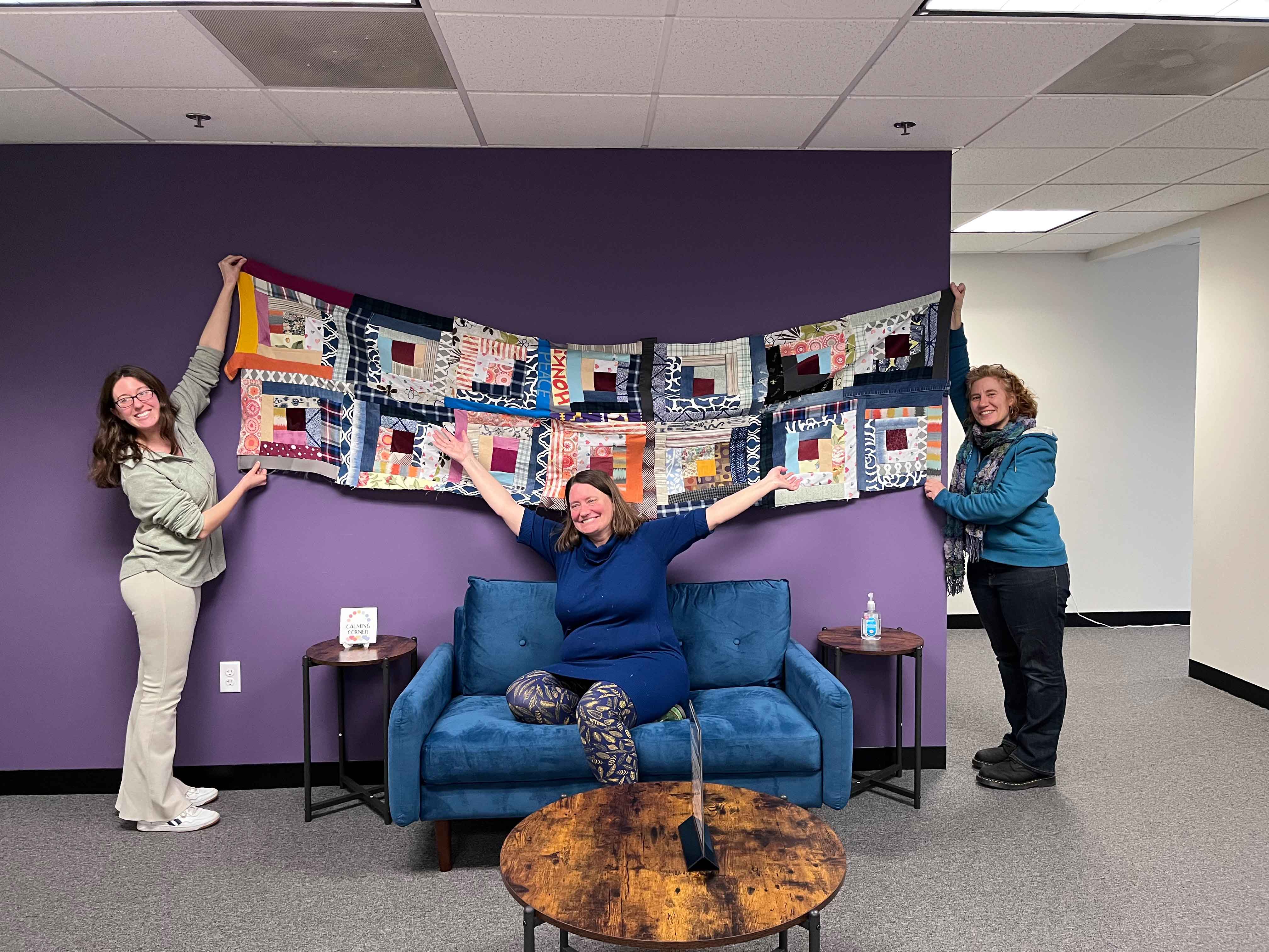 Three women displaying a colorful patchwork quilt above a blue couch in a purple-walled room.