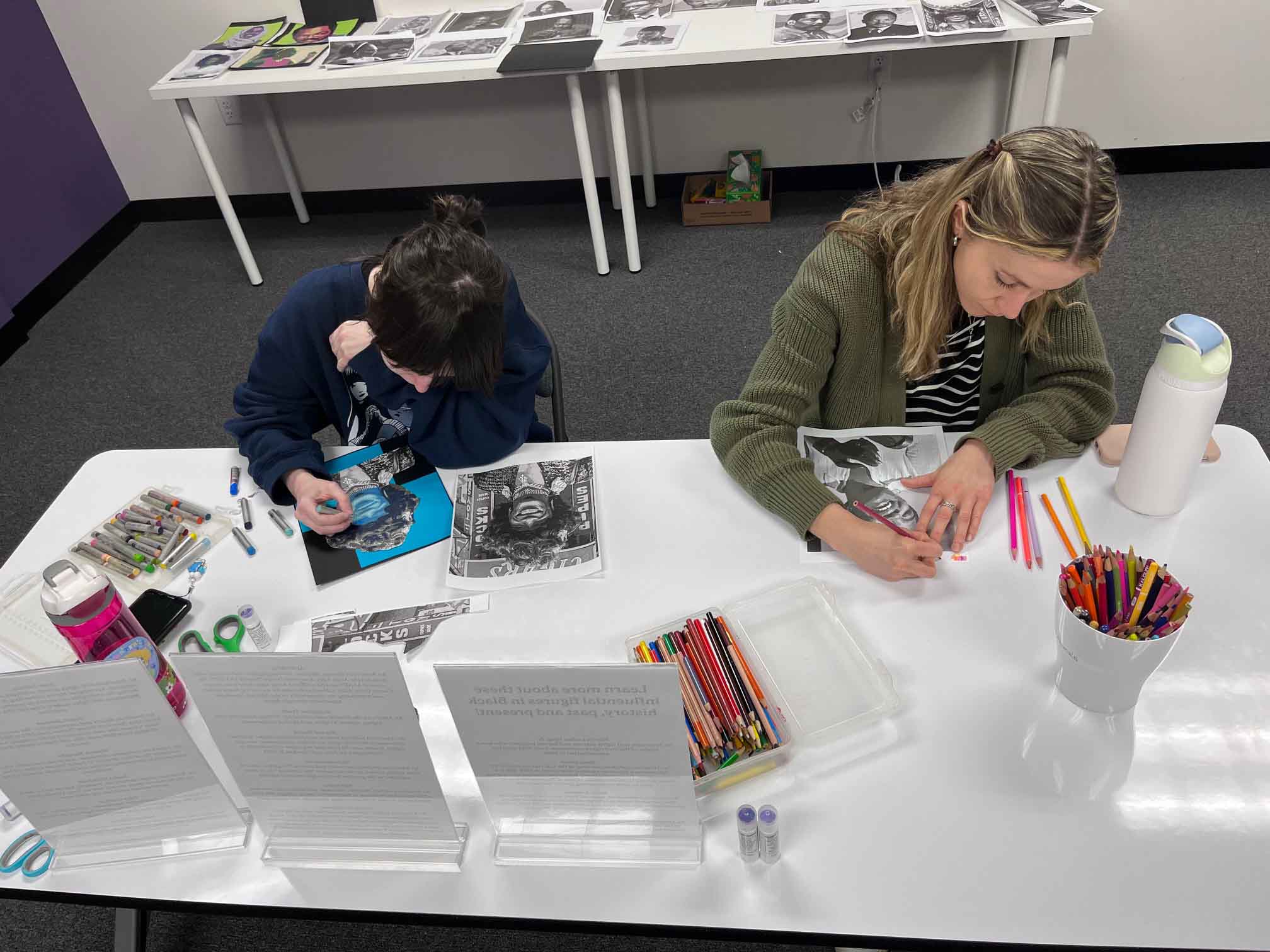 Two women sitting at a table coloring and drawing on portraits with colored pencils and markers.