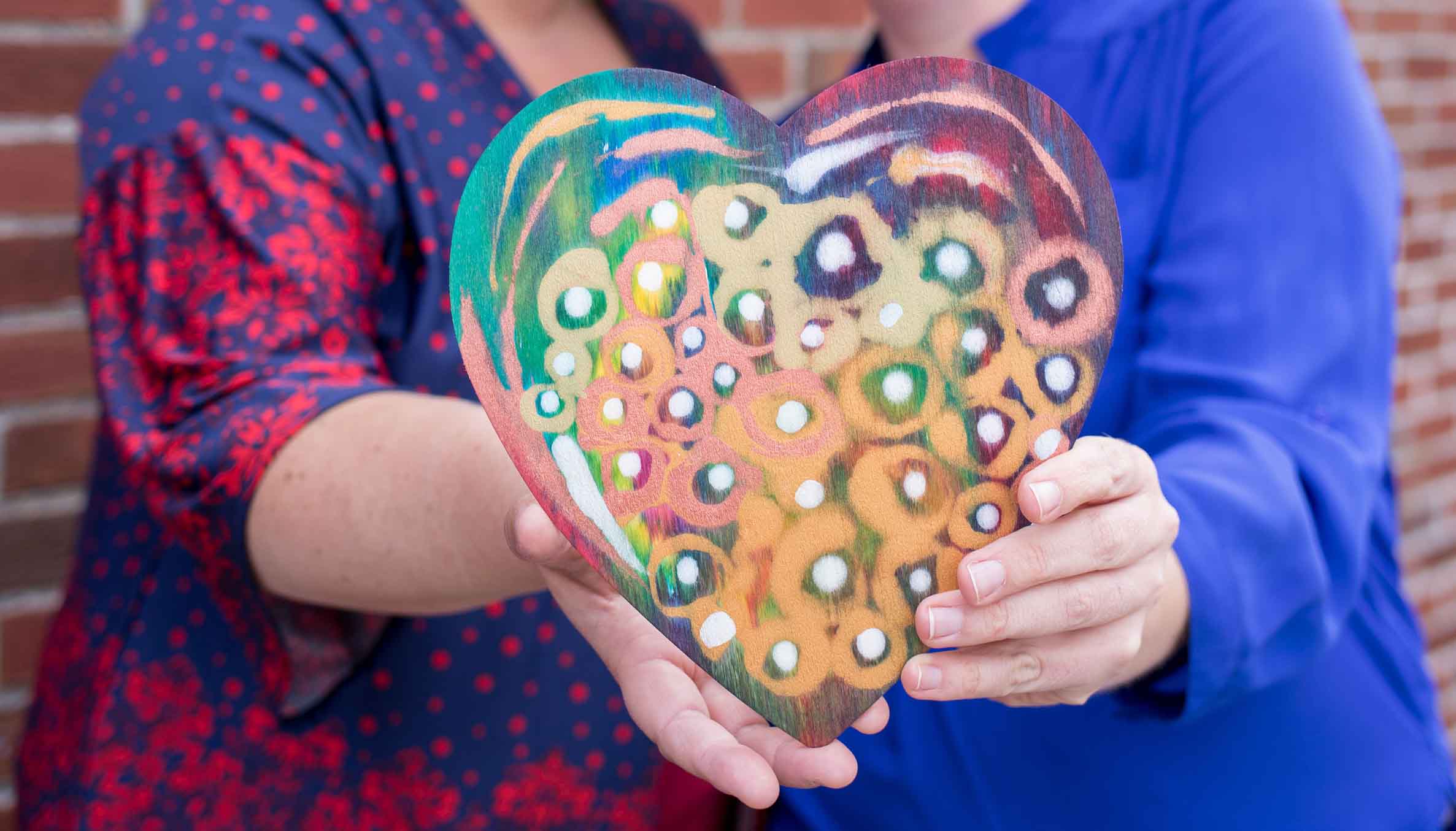 Two people holding a colorful, hand-painted wooden heart with abstract designs.