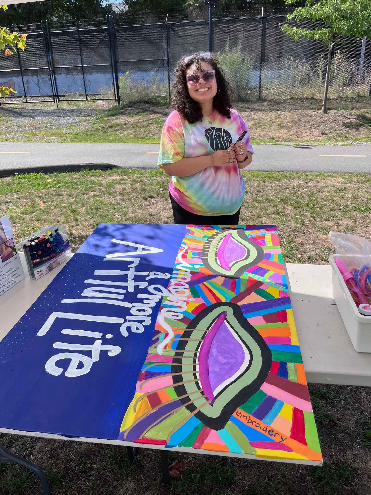 Smiling person in a tie-dye shirt stands behind a table with colorful art and text 'Imagine & groove Artful Life'.