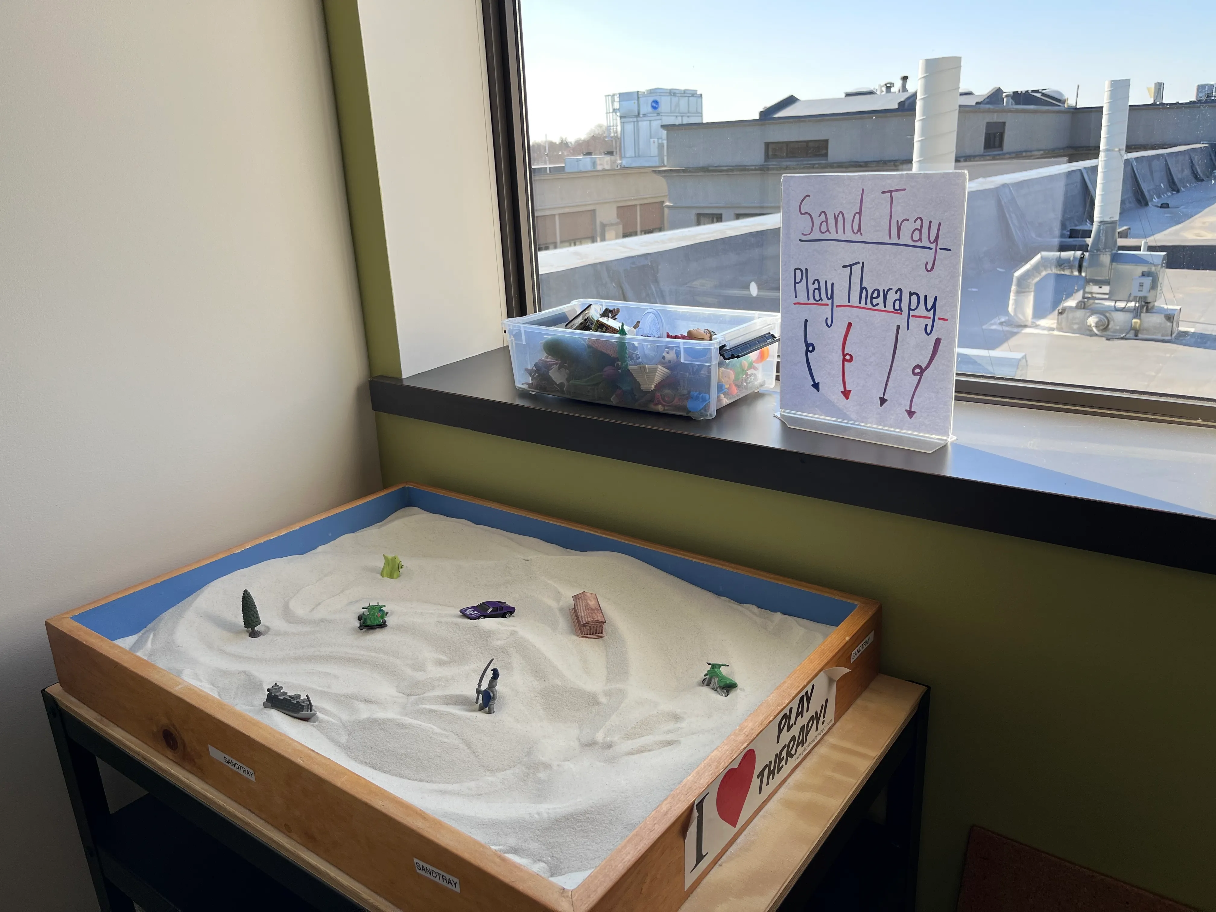Wooden sand tray with small toys and a sign reading 'Sand Tray Play Therapy' by a window.