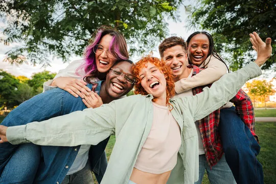Group of five diverse young friends embracing and smiling outdoors in a park under trees.