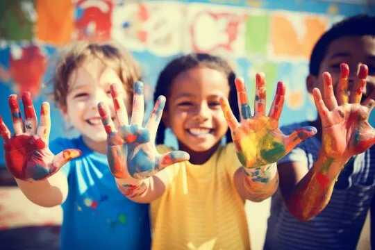 Three happy children showing their hands covered in colorful paint, smiling and standing close together.