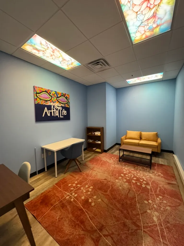 Small room with blue walls, yellow couch, wooden coffee table, desks, colorful art, and red patterned rug.