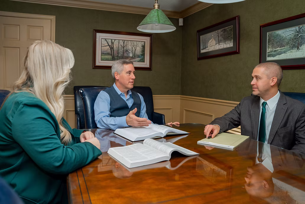 Three people in business attire having a discussion at a conference table with open books.