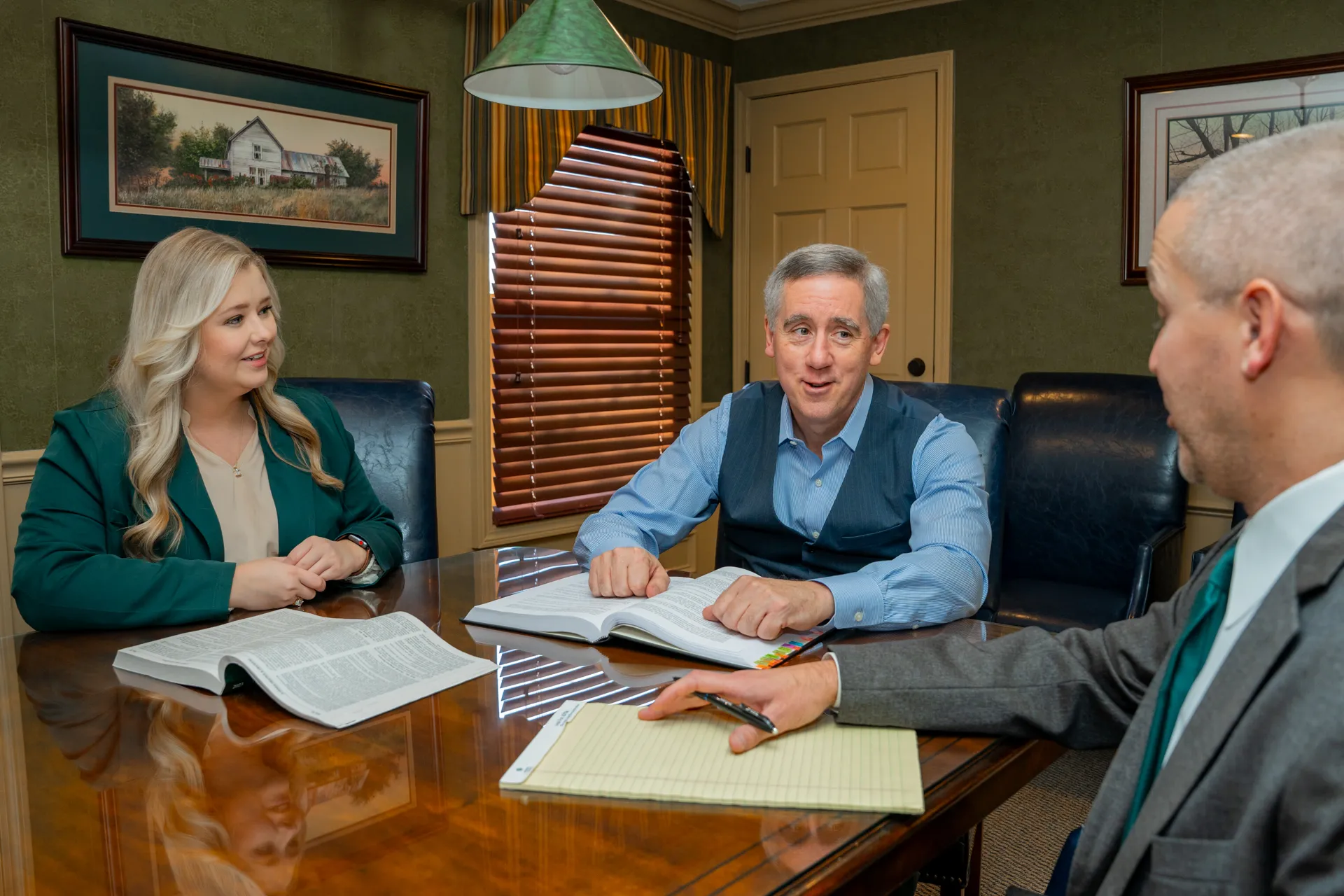 Three professionals seated around a conference table with open books and a notepad in an office.