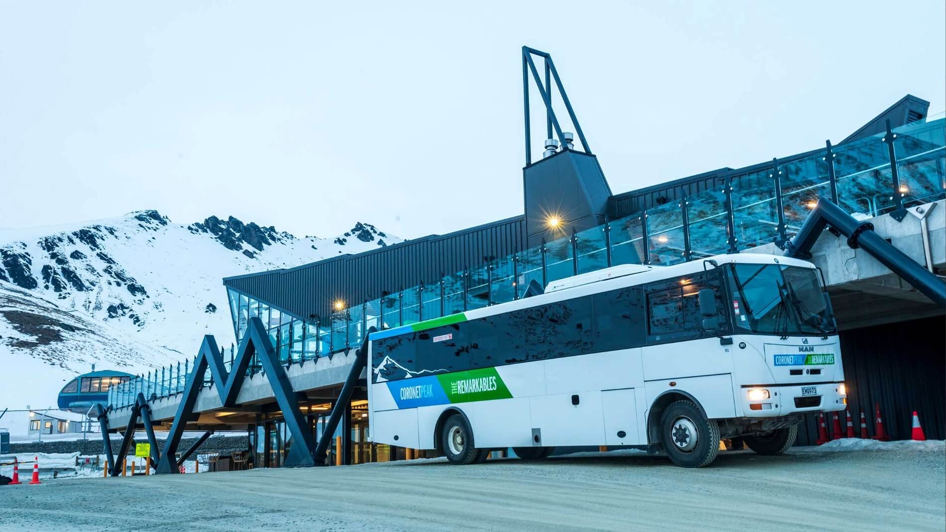 White Coronet Peak ski bus parked beside a modern base building with snowy mountains in the background.