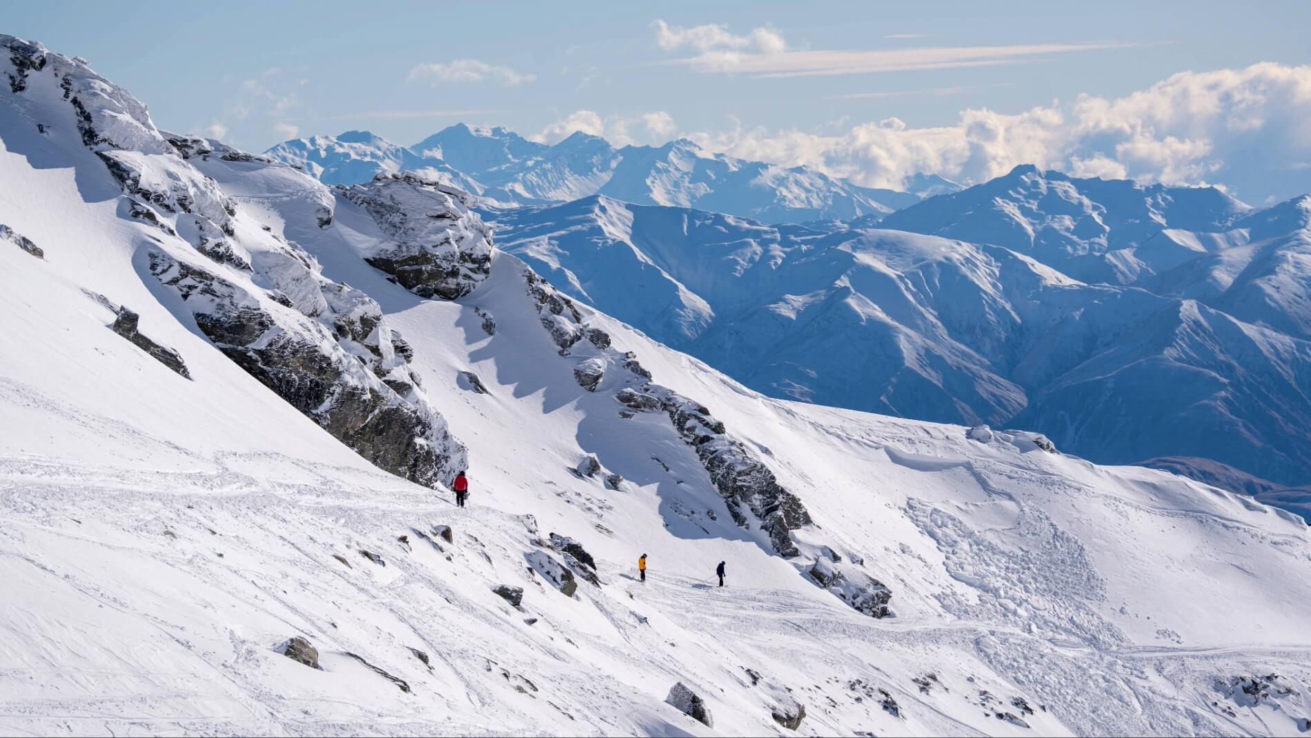 Three skiers traversing a snowy alpine ridge with rugged snow-covered mountains in the background.