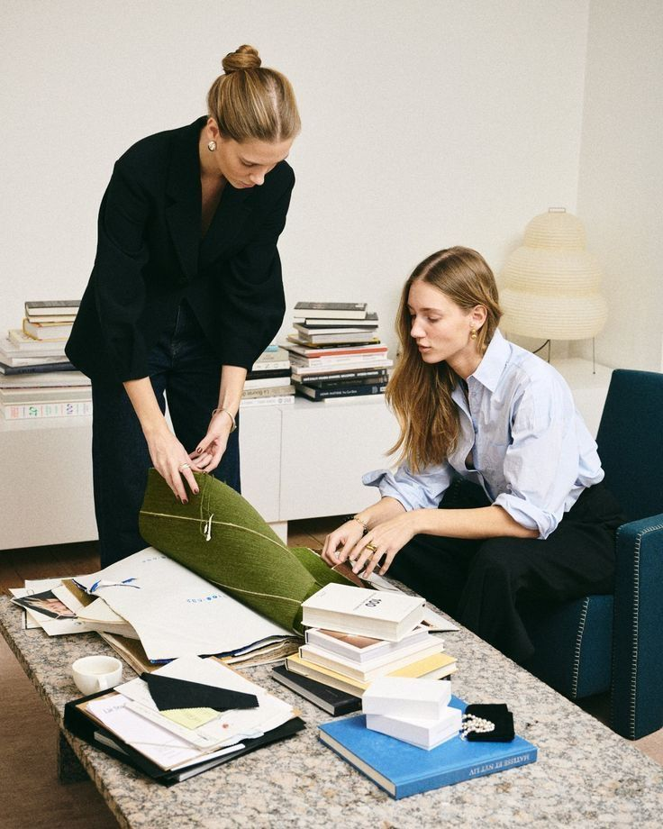 Two women collaborate over fabric samples and books on a marble coffee table in a modern room.