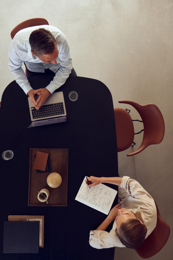 Overhead view of a man and a woman sitting at a black table during a meeting or discussion; the man is using a laptop and the woman is writing notes on a paper.