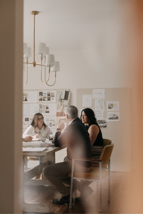 Three people seated around a wooden table in a room with design boards on the wall, engaged in meeting.