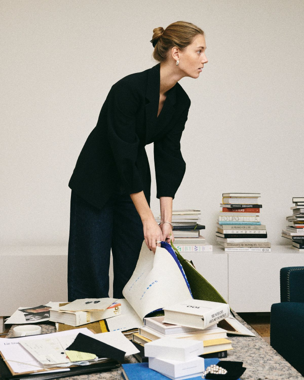 A woman is leaning over a table covered with books, papers, and notebooks.
