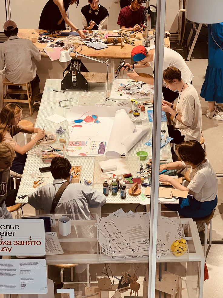 Top view of a workshop with multiple people sitting around a long table engaged in various crafts and art projects, surrounded by tools, paint bottles, papers, and materials.