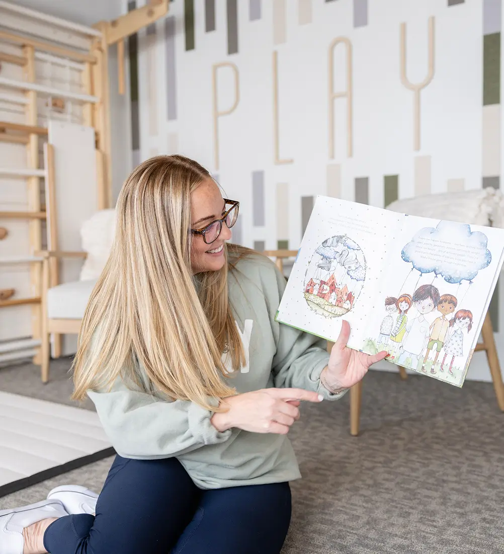 Therapist with long blonde hair reading a children's book in front of the white-and-sage 'PLAY' wall mural at Playful Pathways