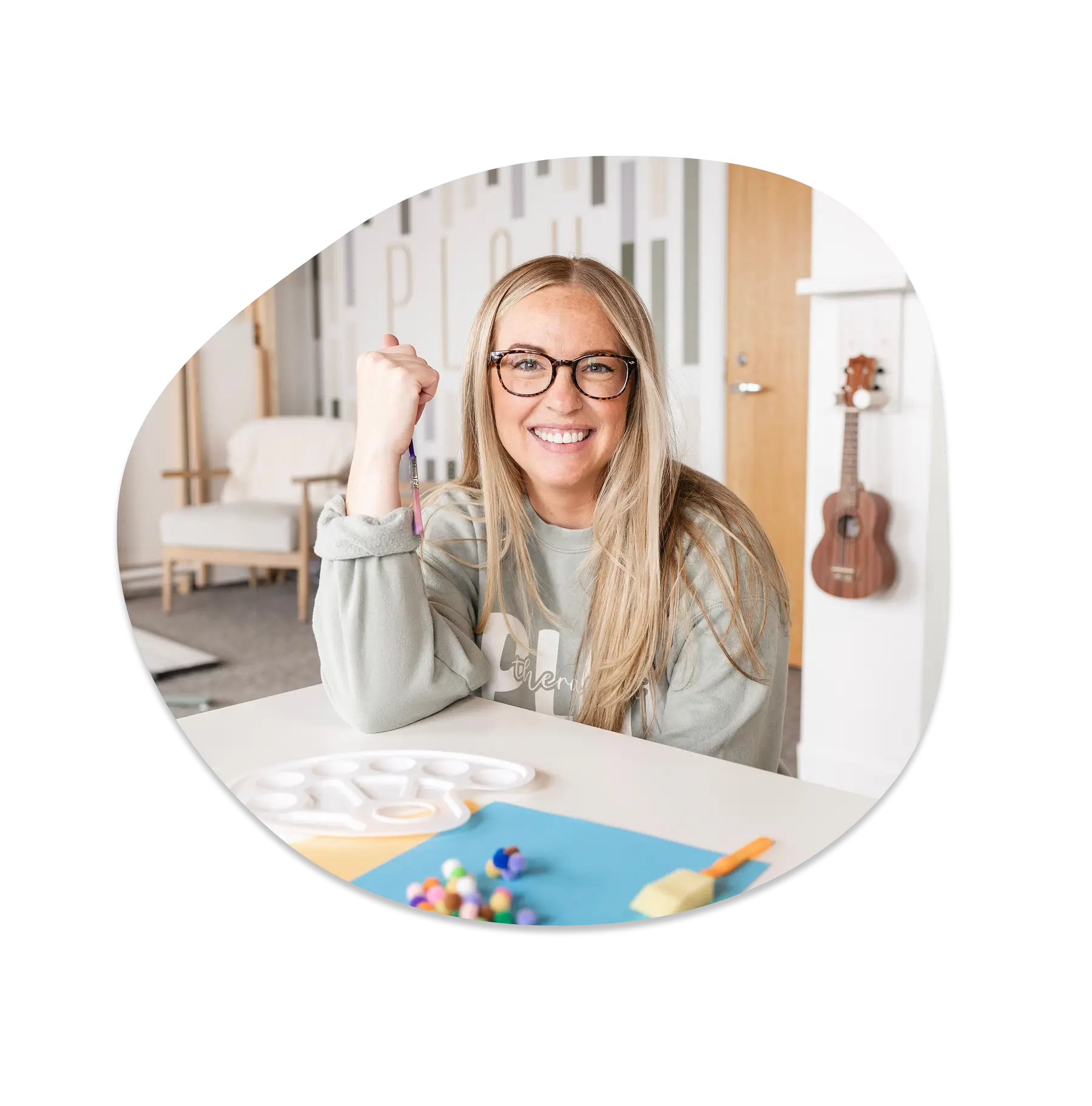 Emilee Greenman, LMSW-C — Therapist at Playful Pathways smiling at an art table holding a paintbrush, with the 'PLAY' mural and a ukulele behind her