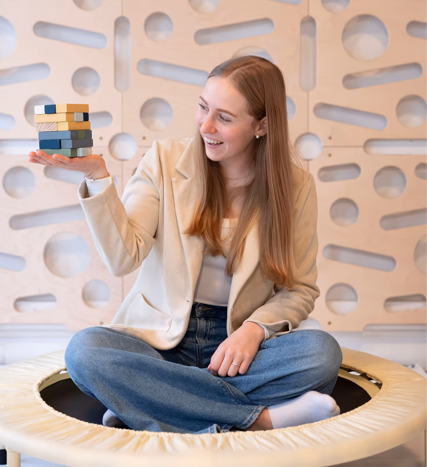 Child therapist at Playful Pathways sitting on a therapy trampoline holding stacking blocks in front of the sensory peg wall