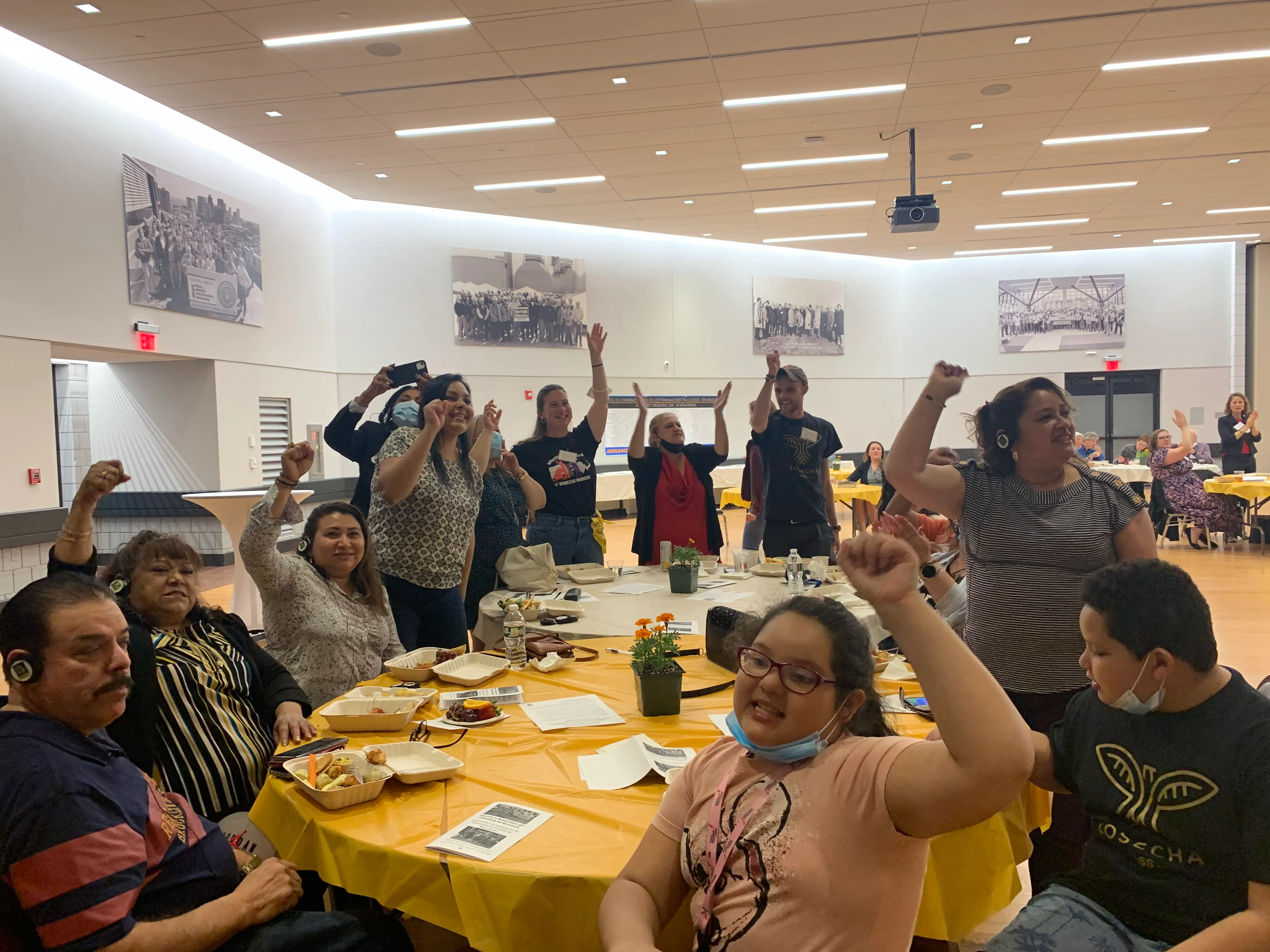People sitting and standing around tables with yellow tablecloths raising their fists in a celebratory or supportive gesture in a well-lit room.