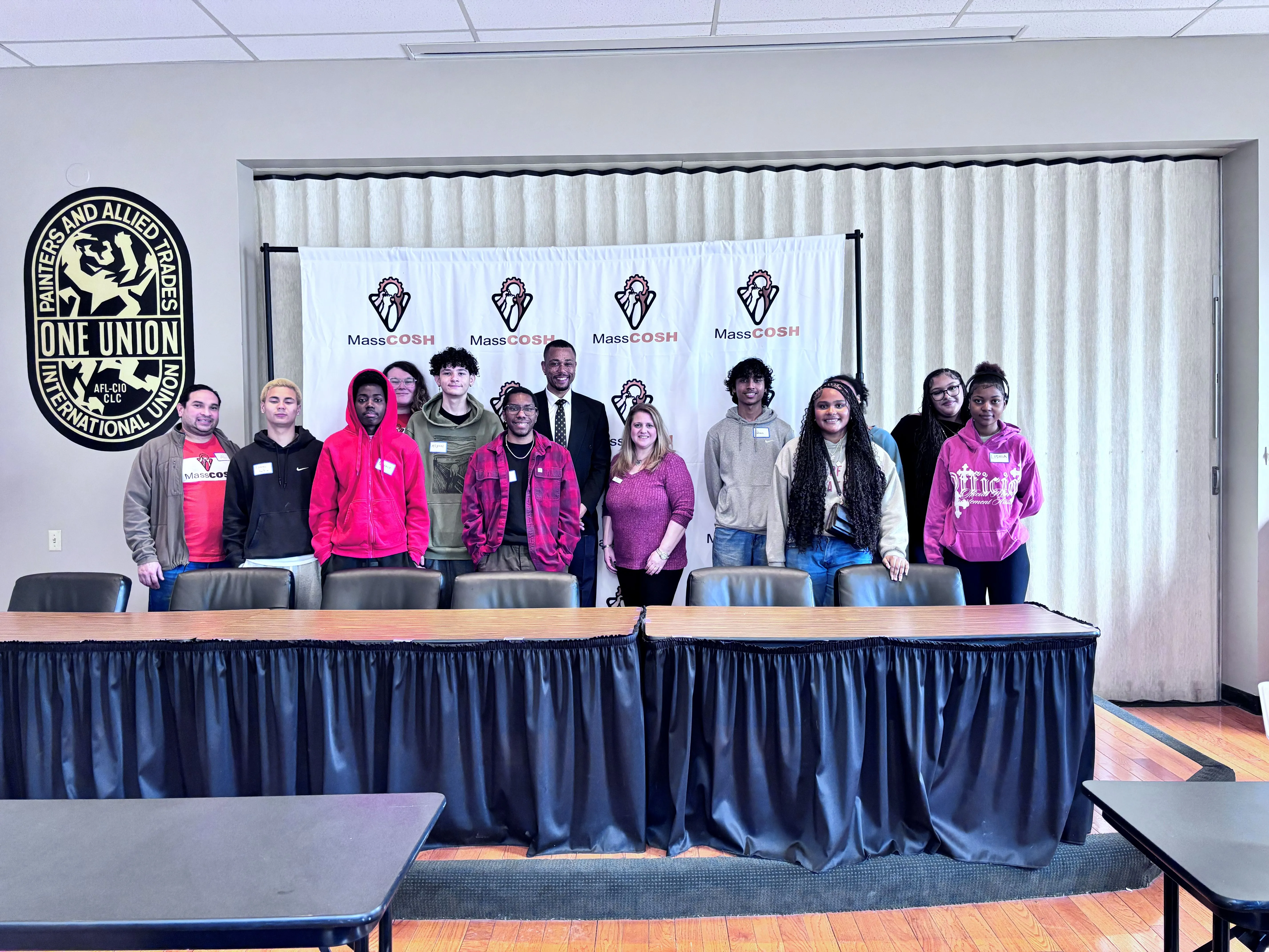 Group of diverse young people and adults standing behind a table with a MassCOSH banner and a Painters and Allied Trades union logo on the wall.