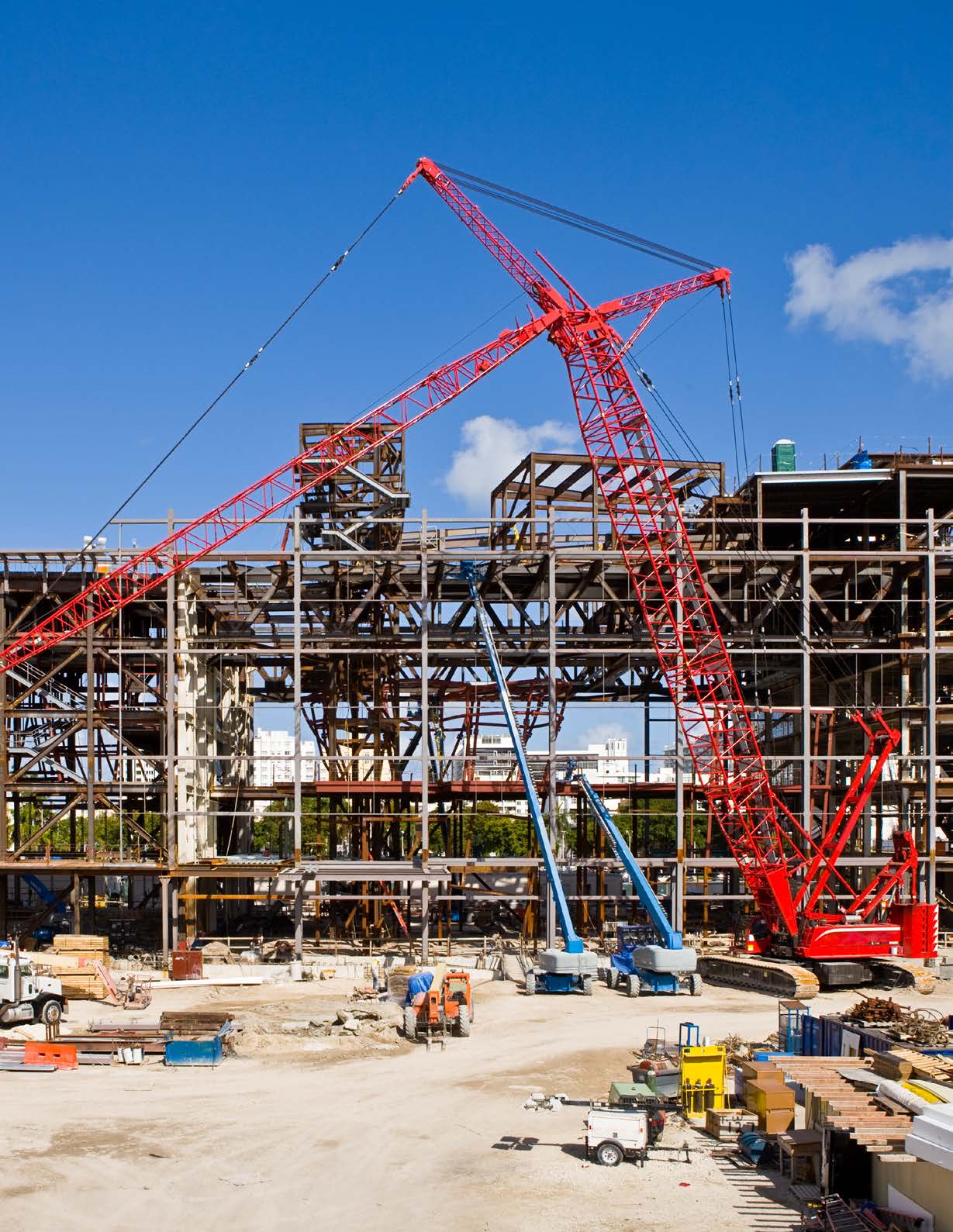 Large commercial construction site with a multi-story steel building frame and a red lattice-boom crane overhead. Blue boom lifts, an orange telehandler, and scattered materials sit on the dirt ground beneath a clear blue sky.