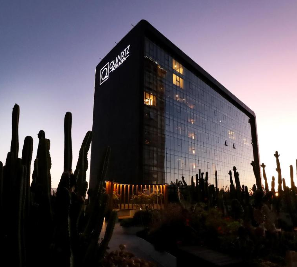 Edificio de Quartz Hotel & Spa con fachada de vidrio reflejando el atardecer, rodeado de cactus.