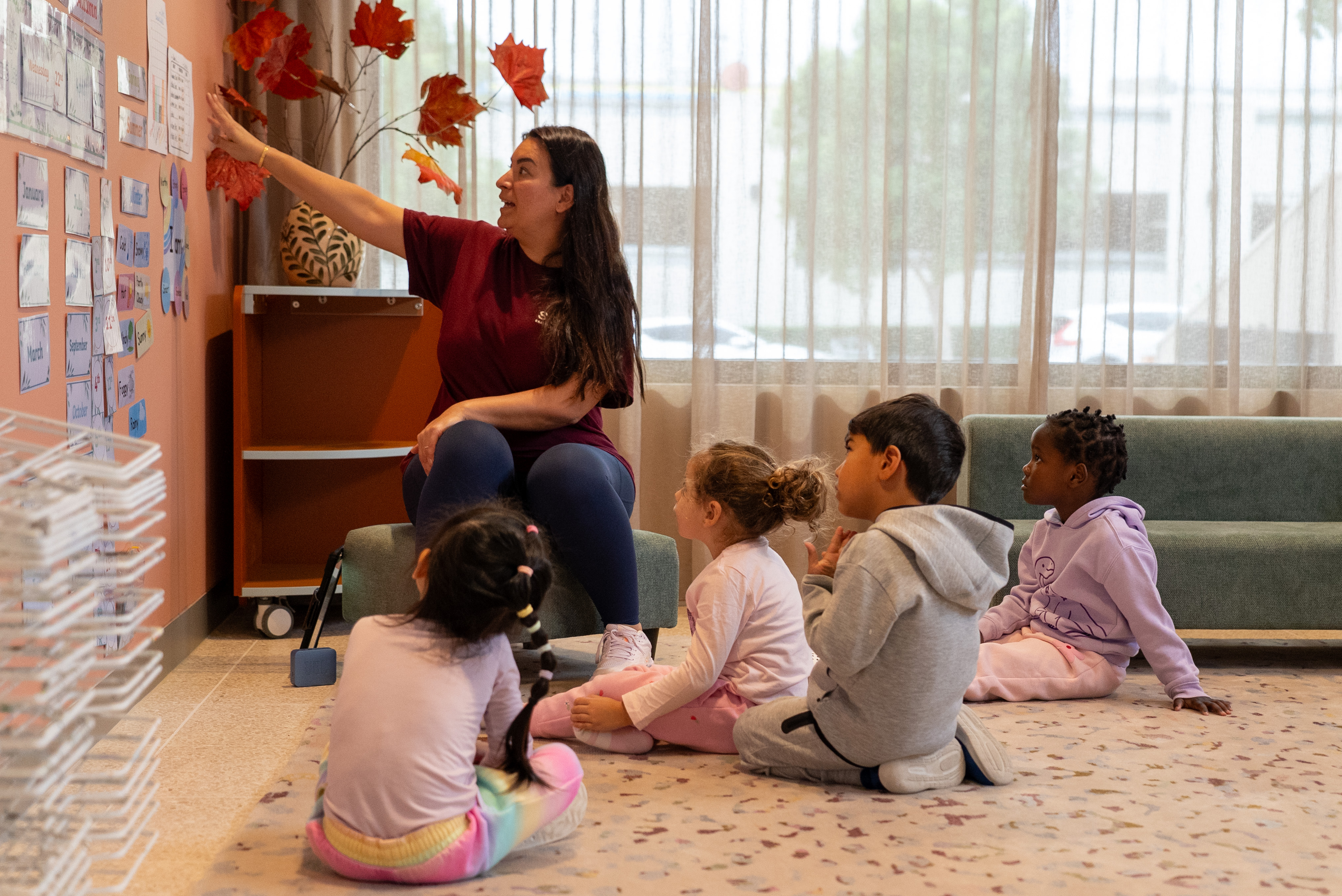 Teacher pointing at a wall chart while four young children sit on the floor watching attentively.