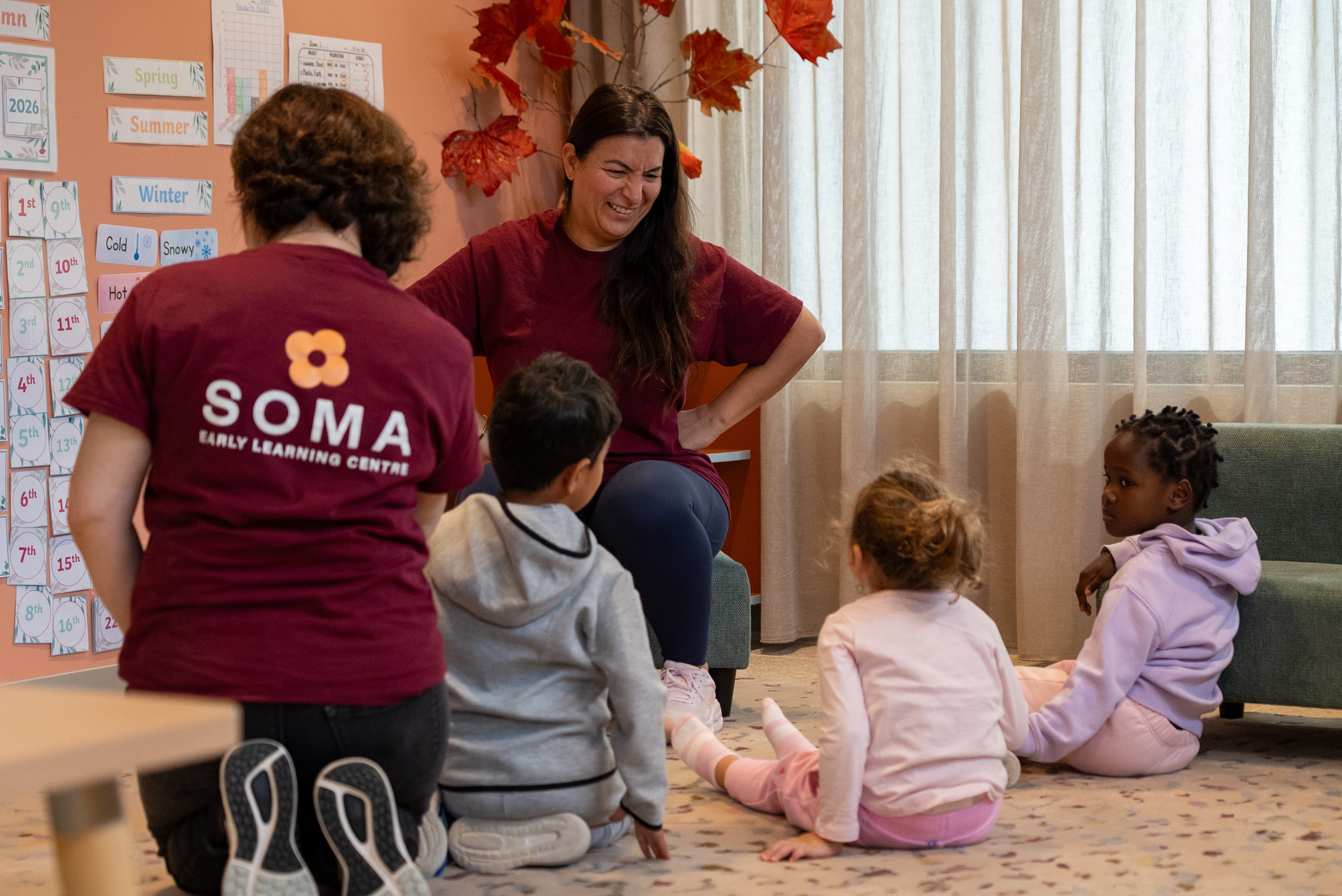 Teachers in maroon shirts with three children sitting on the floor in a colorful classroom corner.