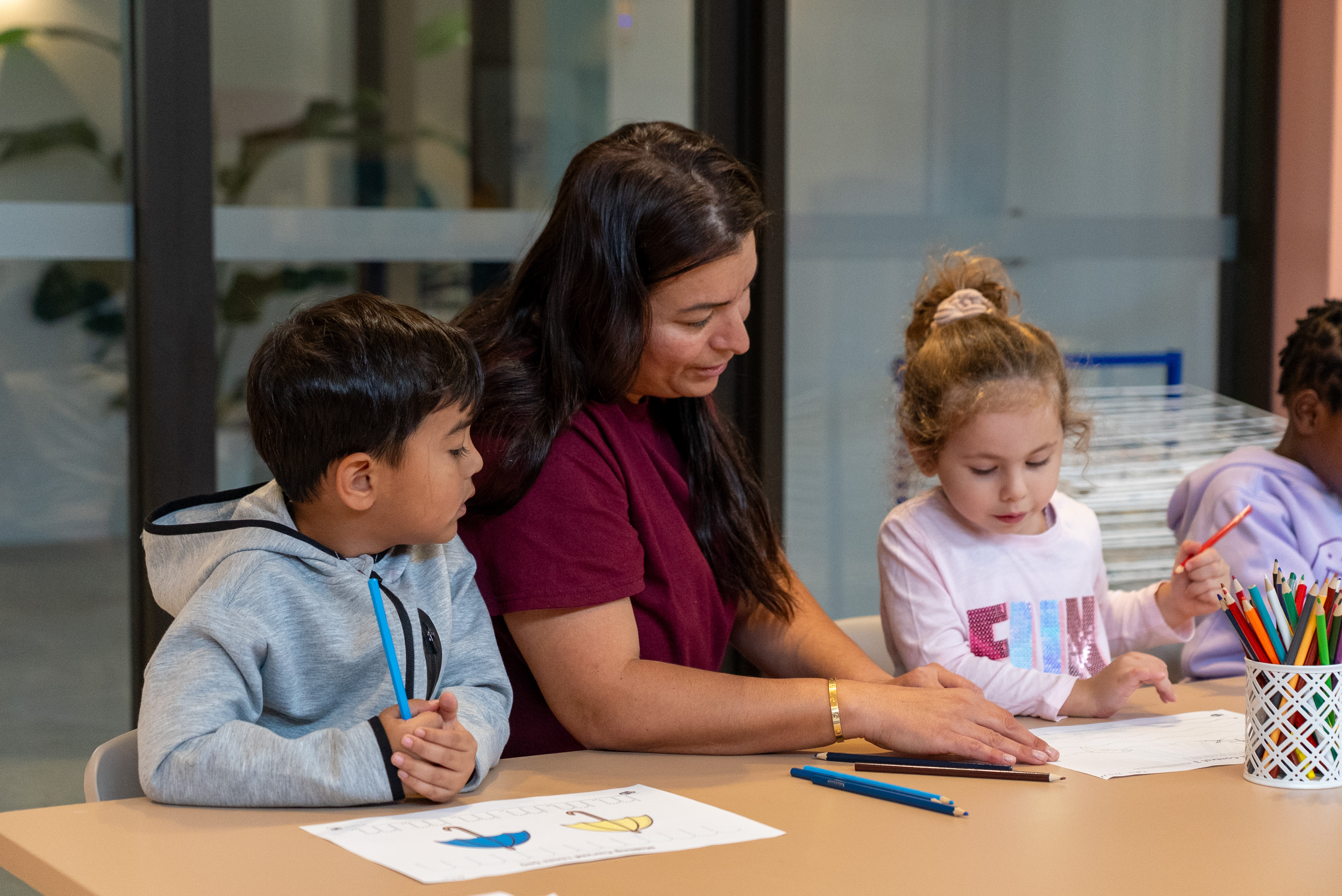 Adult woman helps two children with coloring and writing activities at a classroom table.