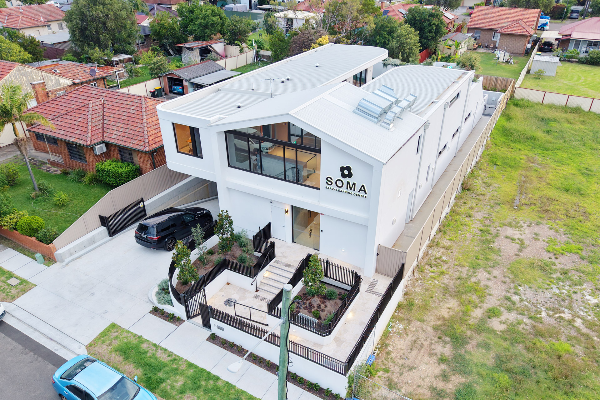 Modern white two-story building labeled SOMA Early Learning Centre with gated entrance and cars outside.