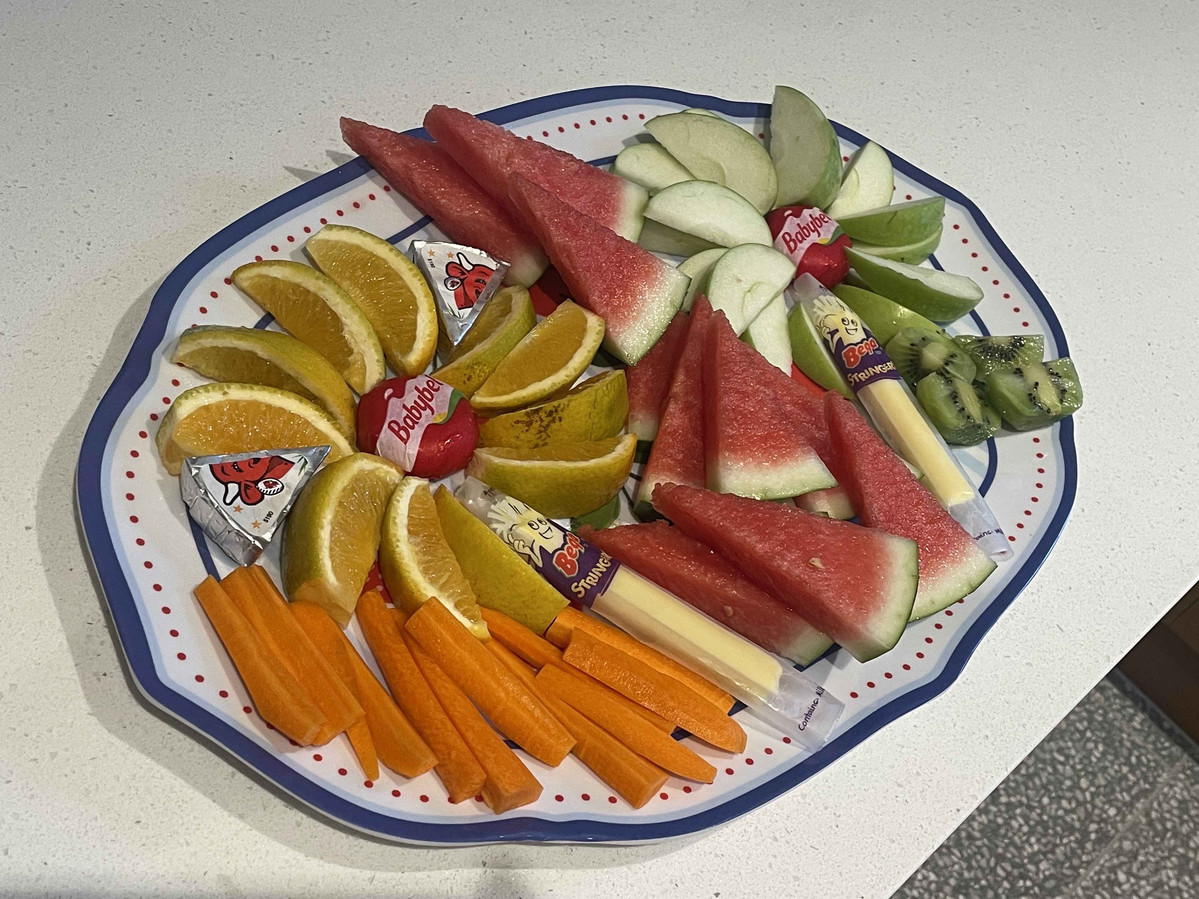 Plate with sliced watermelon, green apple, kiwi, carrots, orange, and cheese snacks.